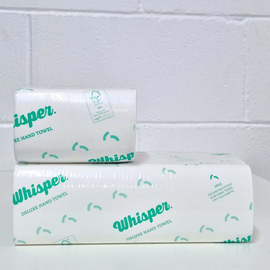 Whisper 3848, Duluxe paper hand towel, Half Size Multifold Slim Hand Towel 1PLY, clear wrapper, green whisper logo and text on packaging, white floor and white brick background background, small stacked on top of a full size