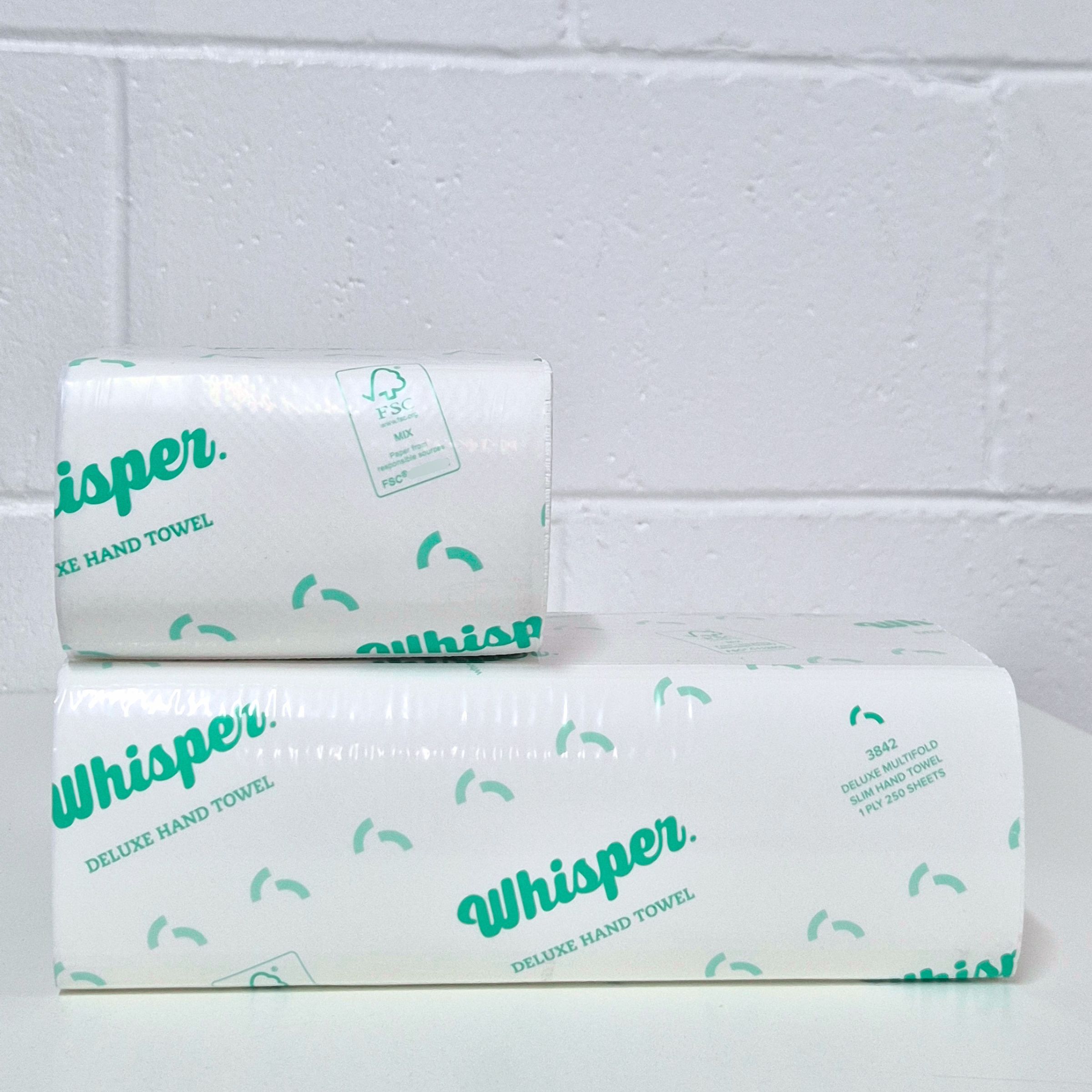 Whisper 3848, Duluxe paper hand towel, Half Size Multifold Slim Hand Towel 1PLY, clear wrapper, green whisper logo and text on packaging, white floor and white brick background background, small stacked on top of a full size