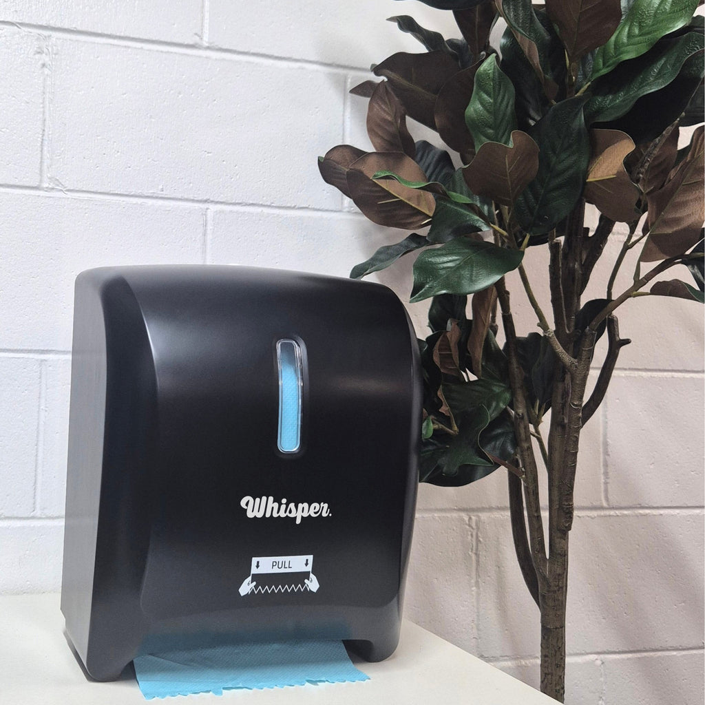 MK8015, Whisper Autocut dispenser, black, white whisper logo, blue paper, roll hand paper towel, on white table, whote brick wall, plant to the right