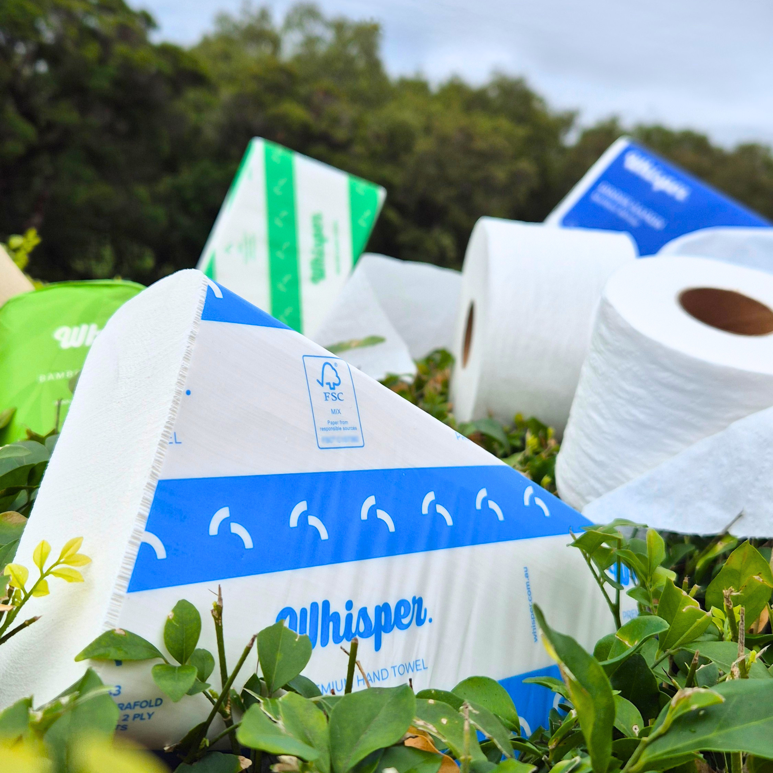 Whisper products with leaves, trees and grey sky in background, whisper blue and white stripped folded hand towel in the front with toilet tissue roll behind, blurry napkins in the background